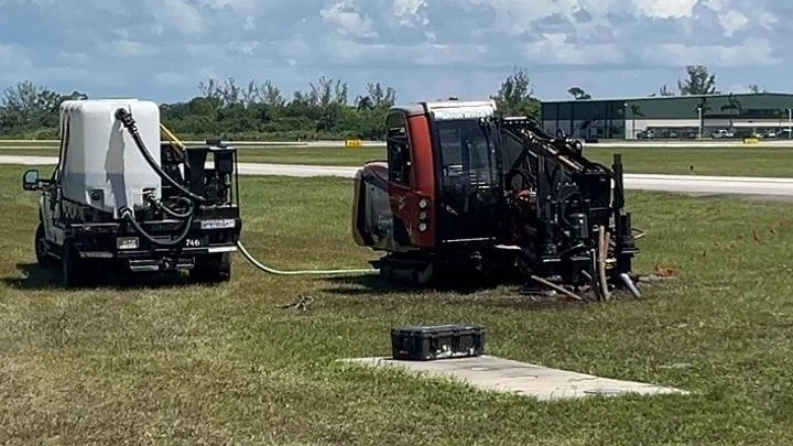 Directional drill with a water-tank support vehicle feeding it by hose, set up on grass beside an airfield runway