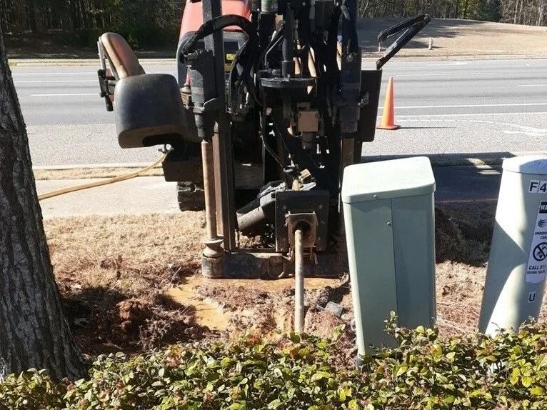 Compact directional drill working between a tree trunk and green utility pedestals beside a road, the kind of tight site where an HDD contractor's coverage matters