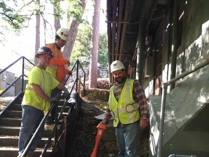 Three-person underground utility contractor crew with a handheld locating instrument at the foot of an outdoor stairway beside a building