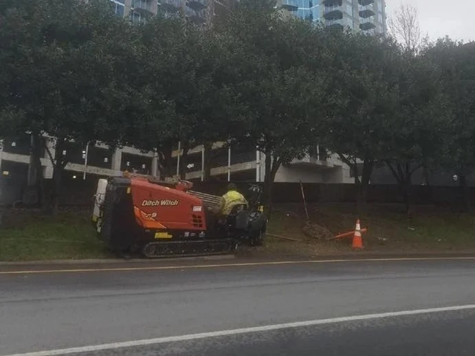Small directional drill working from a grass strip beside a downtown street, one cone marking the bore pit, the trenchless pipe installation method most urban crossings use
