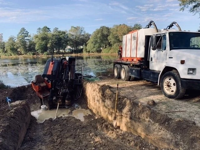 Directional boring rig set in an excavated pit beside a pond with a vacuum truck parked alongside
